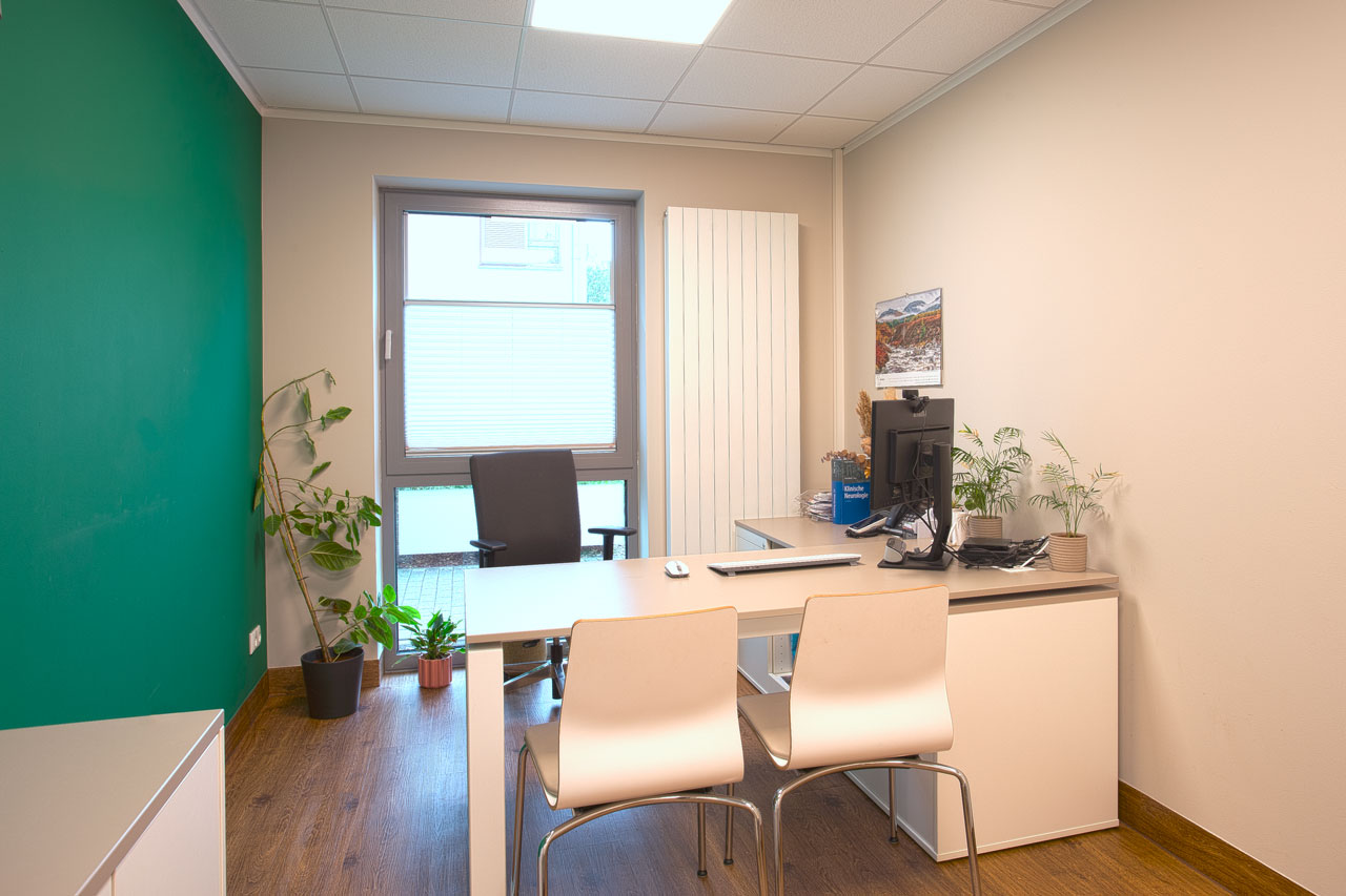 Behandlungsraum mit Schreibtisch, Computer und Stühlen und einer grünen Wand
