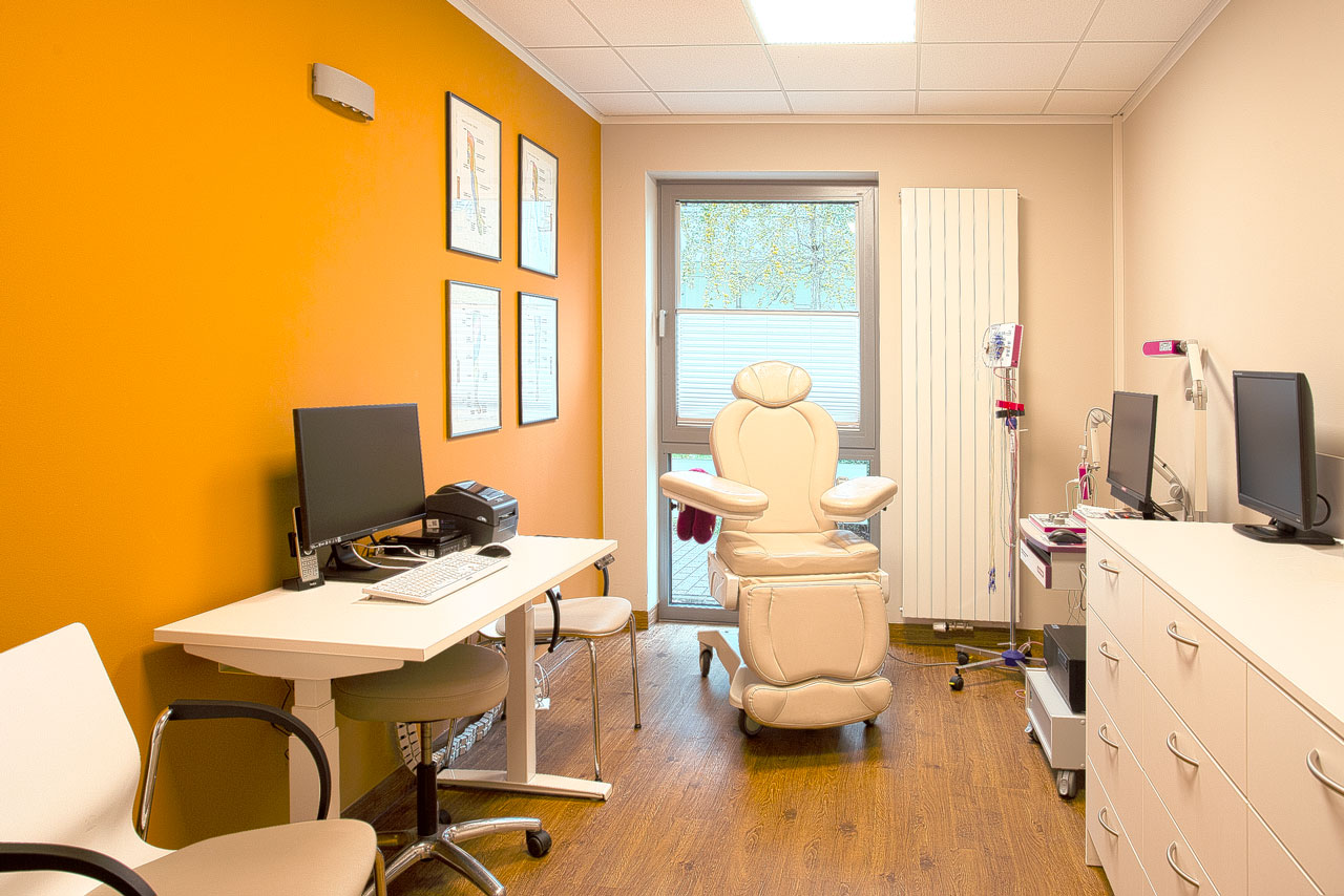 Behandlungsraum mit Schreibtisch, Computer, einem gepolsterten Behandlungsstuhl und einer orangenen Wand